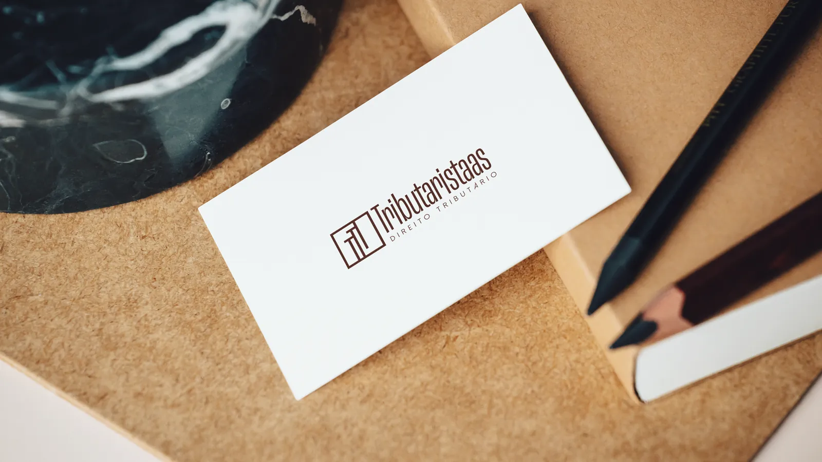 Cartão de visita branco com o logotipo, sobre cortiça, ao lado de lápis
